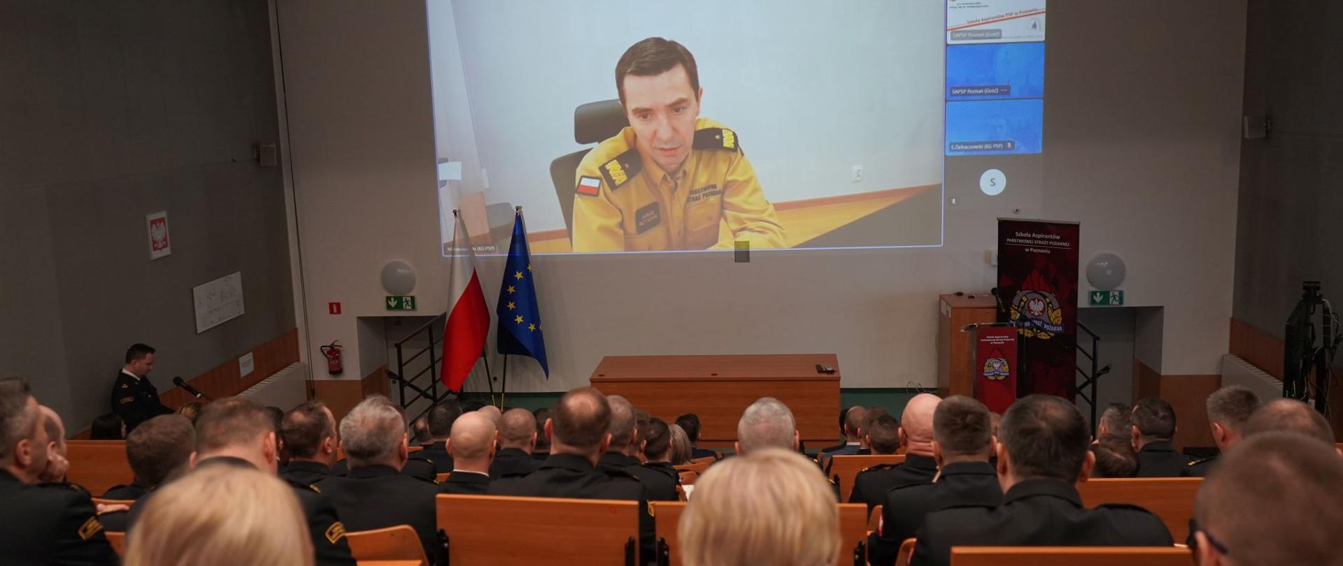 komendant główny PSP za pośrednictwem połączenia wideokonferencyjnego przemawia do uczestników narady zgromadzonych na auli