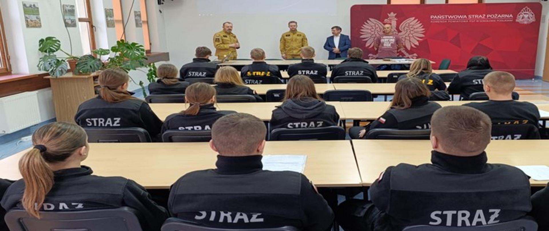 Zdjęcie przedstawia scenę ze spotkania informacyjnego w związku z rozpoczęciem szkolenia podstawowego dla uczniów 4 klasy szkoły ponadposdtawowej.
Pierwszy Plan: Uczestnicy siedzą w rzędach przy jasnych stołach, zwróceni tyłem do obiektywu. Wszyscy ubrani są w czarne, jednolite bluzy/kurtki z dużym, żółtym napisem "STRAŻ" (lub podobnym) na plecach.
Prowadzący: Przy stole prezydialnym na podwyższeniu stoją cztery osoby: dwóch mężczyzn w beżowych/khaki mundurach strażackich, jeden w granatowym garniturze oraz jeden w czerwonej koszulce z nadrukiem (widoczny fragment napisu).
Tło: Za prowadzącymi znajduje się duży, czerwono-biały baner z godłem Polski i napisem "PAŃSTWOWA STRAŻ POŻARNA" oraz informacją o Komendzie Powiatowej PSP. Obok baneru jest duży biały ekran projekcyjny. Po lewej stronie widać okna oraz rośliny doniczkowe.
