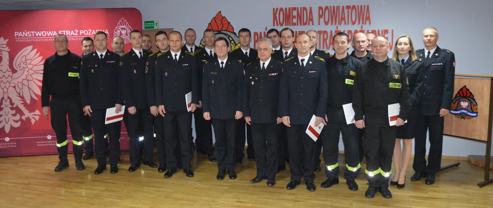 Wręczenie awansów. Świetlica Komendy Powiatowej PSP w Rawiczu. Na tle ścianki reklamowej oraz czerwonego napisu Komenda Powiatowa PSP w Rawiczu do grupowego zdjęcia ustawieni są wszyscy uczestnicy uroczystej zbiórki, tj. awansowani, kierownictwo komendy oraz pozostali strażacy. Występują w umundurowaniu wyjściowym i koszarowym. Awansowani trzymają w ręce biało-czerwone teczki.