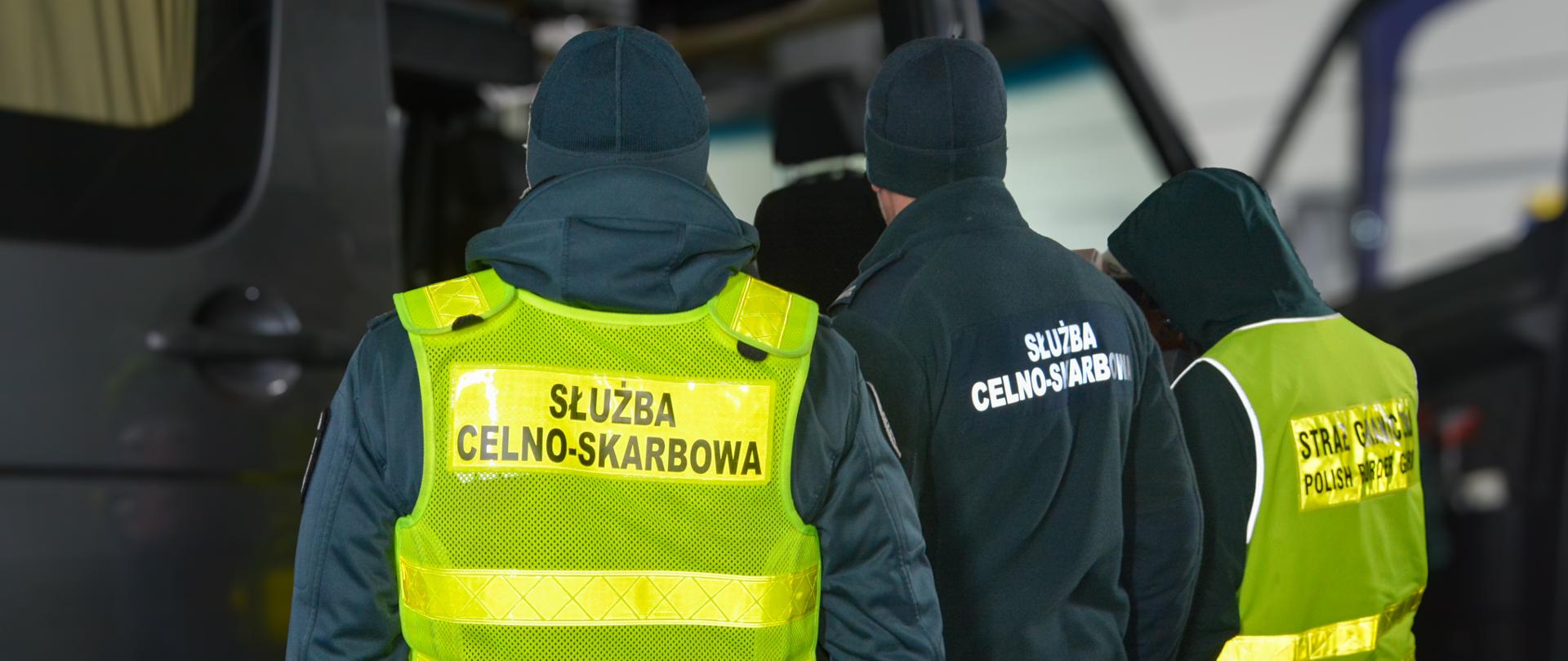 Kilka osób w odzieży służbowej stoi tyłem przy otwartym pojeździe, patrząc do jego wnętrza.