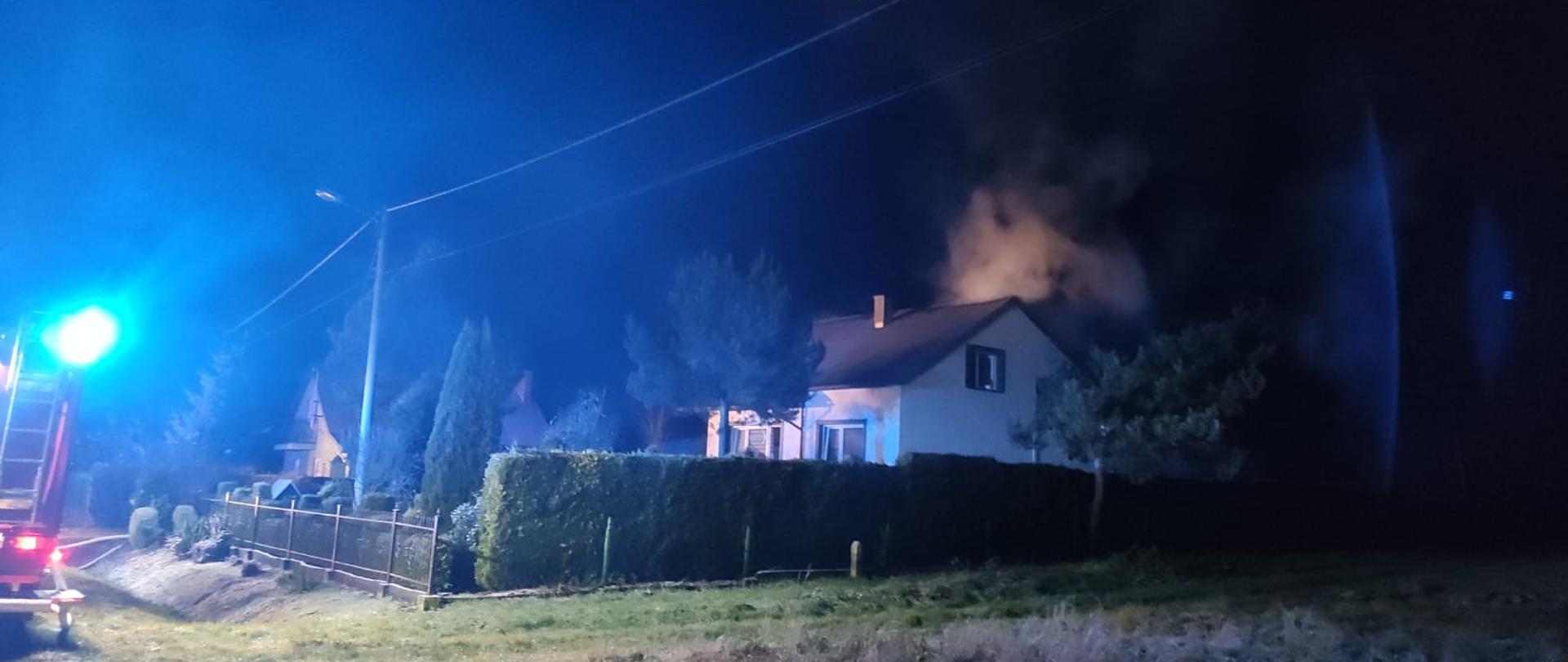 Zdjęcie przedstawia dom jednorodzinny w porze nocnej. Z nad budynku widać unoszące kłęby białego dymu.