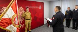 Strażak Państwowej Straży Pożarnej w ubraniu służbowym na tle banneru koloru czerwonego z napisem Państwowa Straż Pożarna i Komenda Wojewódzka PSP w Opolu - str. Kamil Buła w trakcie ślubowania. Treść ślubowania czyta nadbryg. Krzysztof Kędryk Opolski Komendant Wojewódzki Państwowej Straży Pożarnej. Po prawej stronie Zastępca Opolskiego Komendanta Wojewódzkiego PSP st. bryg. Wojciech Nawara oraz naczelnik wydziału technicznego Komendy Wojewódzkiej PSP w Opolu st. bryg. Ryszard Sałek.
Strażak ślubuje na sztandar Komendy Wojewódzkiej PSP w Opolu widoczny po lewej stronie kadru.