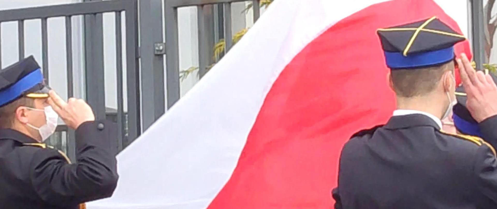 Zdjęcie przedstawia trzech funkcjonariuszy Państwowej Straży Pożarnej. Funkcjonariusze ubrani są w galowe mundury. Dwóch z nich oddaje honory podczas podnoszenia flagi na masz. Trzeci z nich dokonuje wciągnięcia flagi na maszt. Zdjęcie wykonane przed Jednostką Ratowniczo – Gaśniczą numer 2 w Radomiu. 