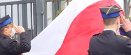Zdjęcie przedstawia trzech funkcjonariuszy Państwowej Straży Pożarnej. Funkcjonariusze ubrani są w galowe mundury. Dwóch z nich oddaje honory podczas podnoszenia flagi na masz. Trzeci z nich dokonuje wciągnięcia flagi na maszt. Zdjęcie wykonane przed Jednostką Ratowniczo – Gaśniczą numer 2 w Radomiu. 