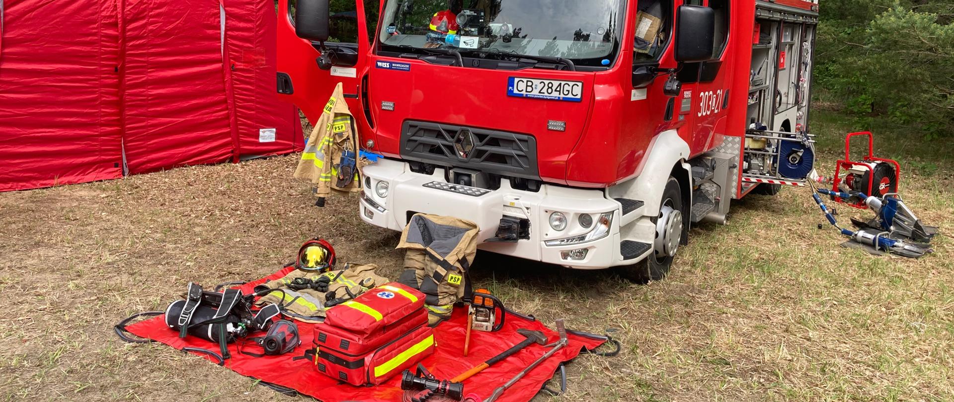 Przód samochodu strażackiego czerwonego stojącego na łące przy lesie. Przed samochodem na czerwonej plandece leży sprzęt ratowniczo-gaśniczy, z lewej przód czerwonego namiotu pneumatycznego. 