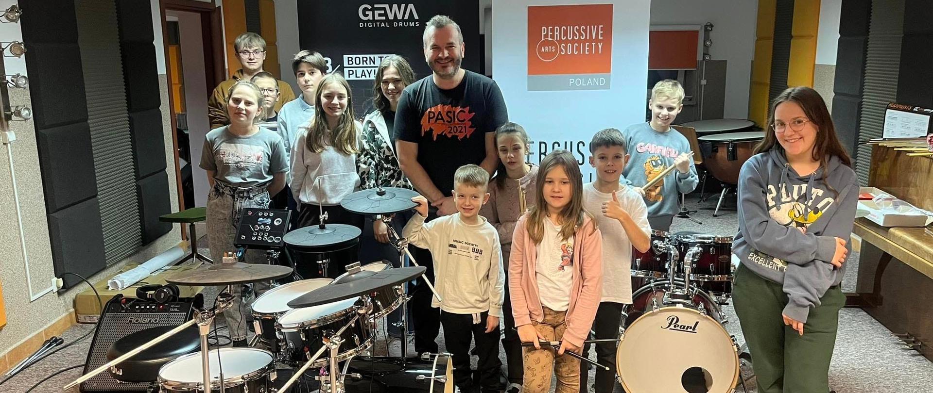Zdjęcie grupowe. 12 uczniów w różnym wieku. Nauczyciel stoi pomiędzy nimi. Po lewej stronie stoi perkusja, po prawej pianino.