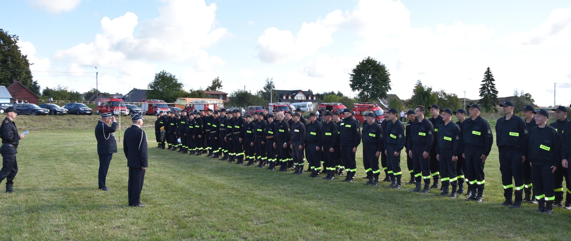 Zdjęcie przedstawia strażaków ustawionych w szyku w tle pojazdy pożarnicze i cywilne