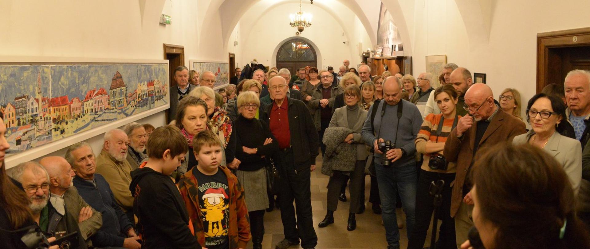 W holu muzeum bardzo liczna grupa ludzi, po lewej stronie widoczne na ścianie dwie długie, poziome kompozycje malarskie