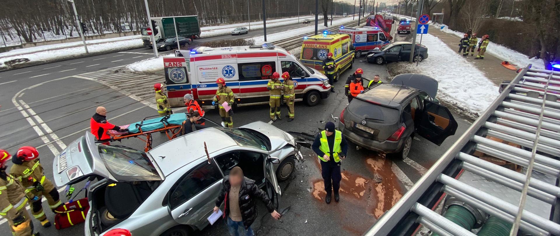 Widok na miejsce zdarzenia, prowadzenie czynności przez funkcjonariuszy.
Na fotografii zauważyć można widok skrzyżowania, na którym doszło do wypadku. Na pierwszym planie znajdują się dwa samochody biorące udział w zderzeniu- srebrna Mazda oraz czarny Nissan. Przy srebrnym samochodzie trwa udzielanie pomocy osobie poszkodowanej, która znajduje się wewnątrz pojazdu przez Zespół Ratownictwa Medycznego. Dalszy plan zdjęcia prezentuje trzeci wrak biorący udział w wypadku oraz 3 karetki pogotowia ratunkowego. Na fotografii również zauważyć można funkcjonariuszy straży pożarnej oraz ich samochody. Zdjęcie zostało wykonane z dachu wozu strażackiego.