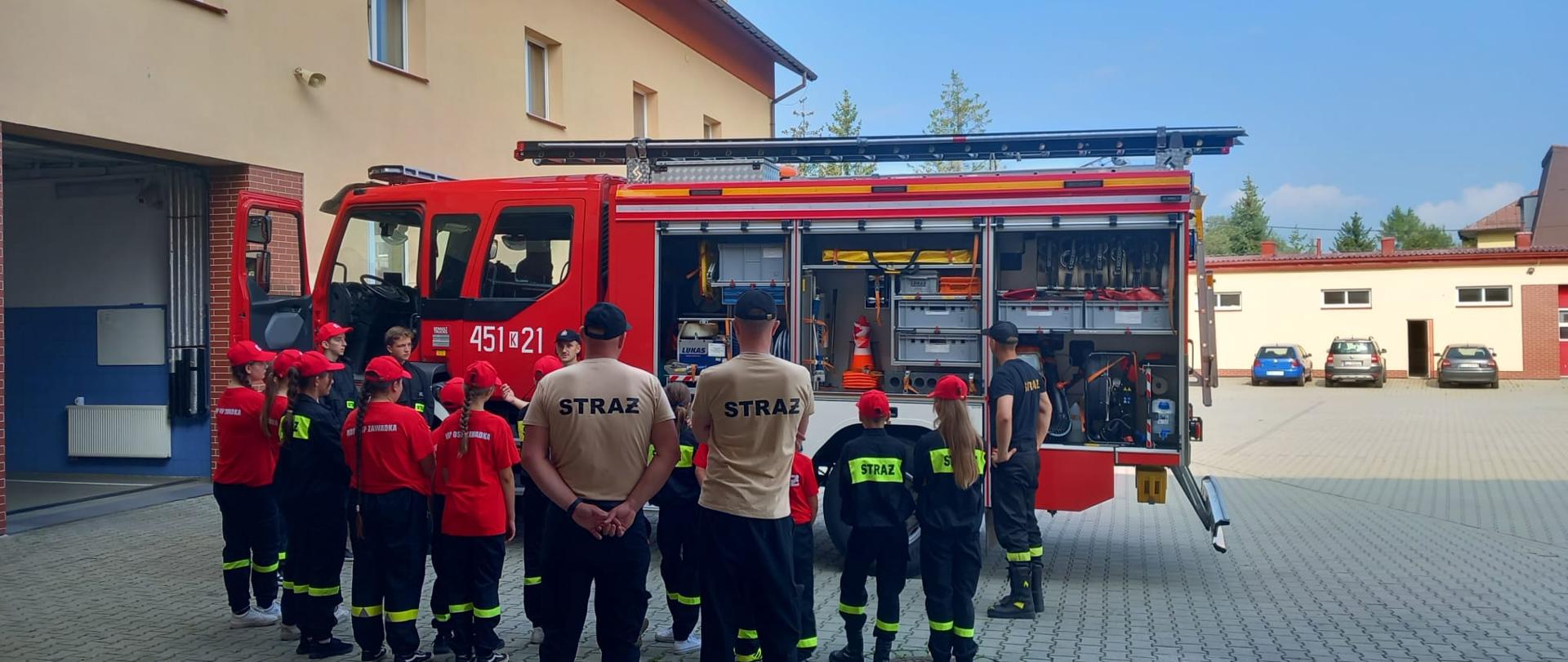 Na tle samochodu strażackiego stoi grupa dzieci wraz z opiekunami w umundurowaniu strażackim