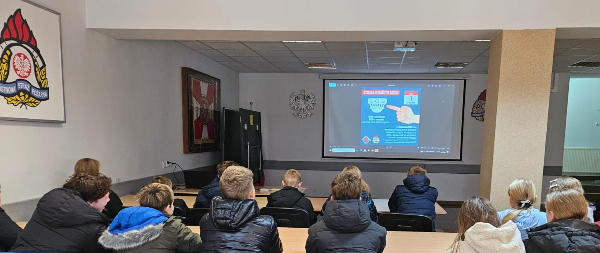Grupa uczniów siedzi w sali wykładowej komendy straży pożarnej i ogląda prezentację na dużym ekranie dotyczącą bezpieczeństwa pożarowego i czujek.