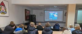 Grupa uczniów siedzi w sali wykładowej komendy straży pożarnej i ogląda prezentację na dużym ekranie dotyczącą bezpieczeństwa pożarowego i czujek.