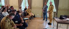 Strażak i mężczyzna po jego prawej stronie stoją przed zgromadzonymi uczestnikami szkolenia i przemawiają do nich. Za nimi widoczne dwa okna z brązowymi zasłonami. 