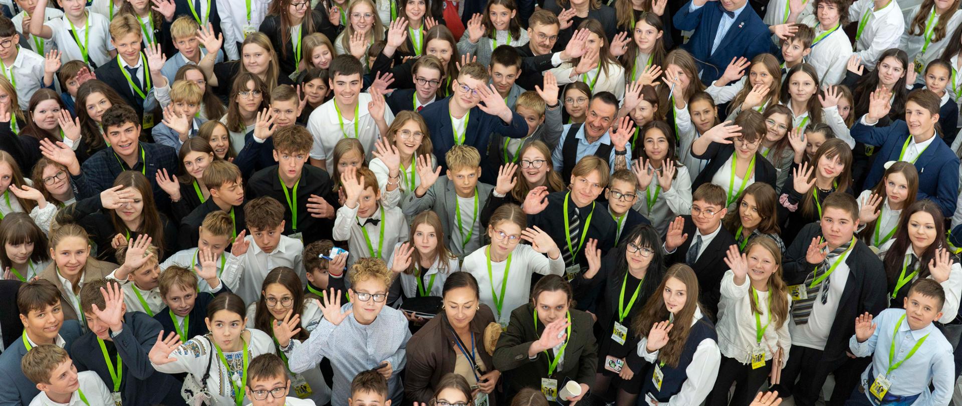 Zdjęcie wykonane z góry. Grupa uczniów i uczennic - członków XXXI sesji Sejmu Dzieci i Młodzieży macha do aparatu. 