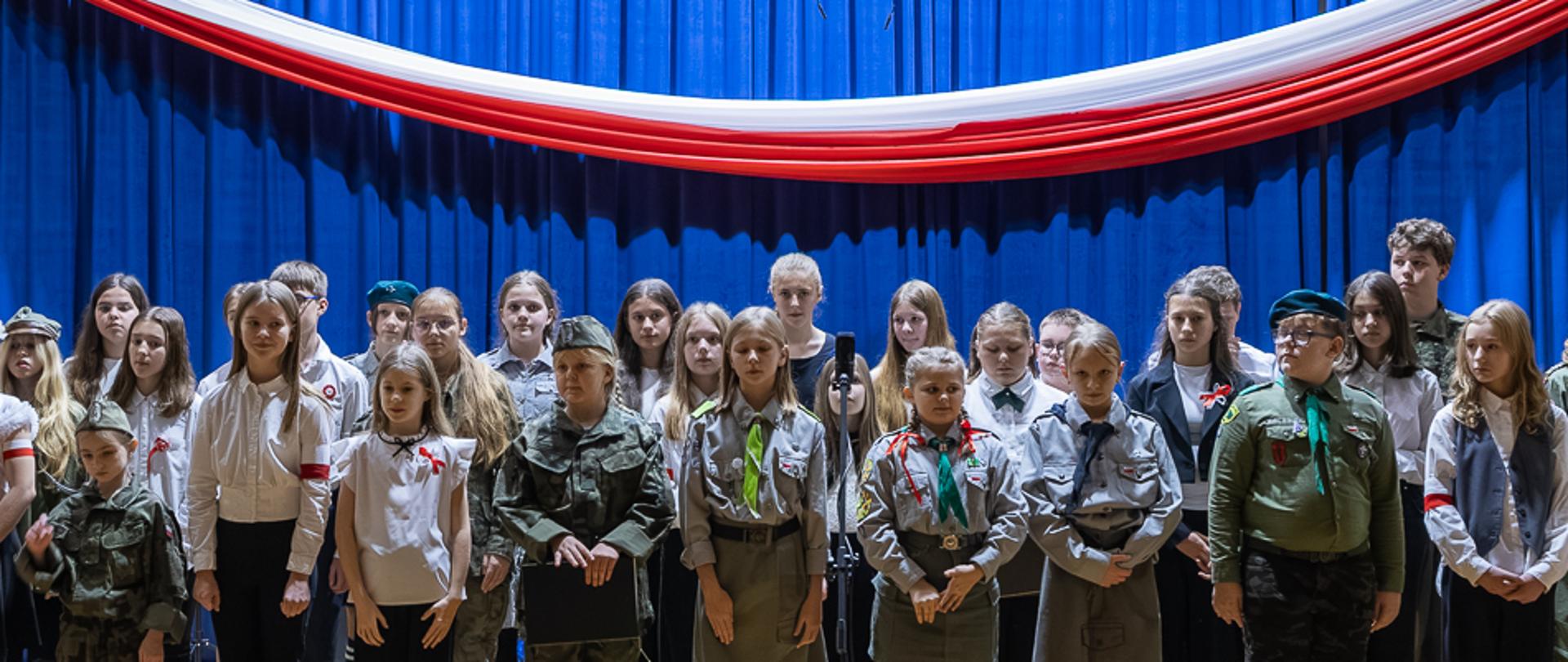 kilkadziesiąt osób stojących na scenie, za nimi niebieska kotara i biało-czerwona wstęga, z przodu publiczność