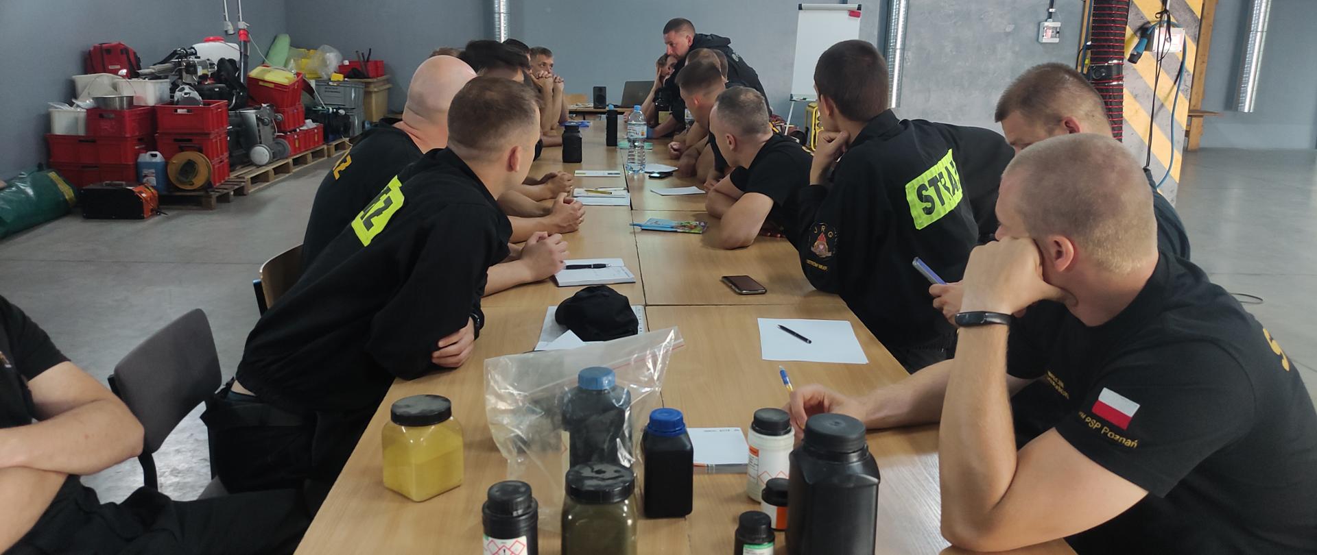 grupa strażaków siedi przy długi mstole , na stole znajdują się środki chemiczne