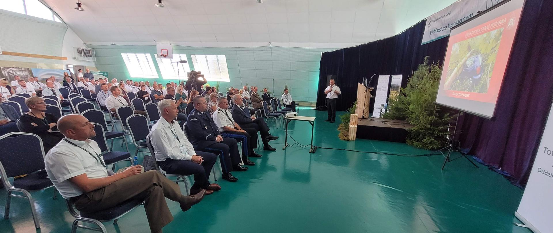 Duże wnętrze sami gimnastycznej z posadzką w kolorze zielonym z białym sufitem. NA sali równo rołżożone krzesełka a na nich siedząca widownia, głównei osoby w garniturach, mundurach i koszulkach w sumie około kilkudziesięciu osób. Po prawej stronie kadru Funkcjonariusz ubrany w białą koszulę z krótkim rękawem referuje pokaz slajdów. Na pierwszym planie rzutnik i ekran na którym wyświetla się pokaz slajdów. Na skraju zdjęcia widoczny rzutnik multimedialny i ekran z pokazem slajdów.