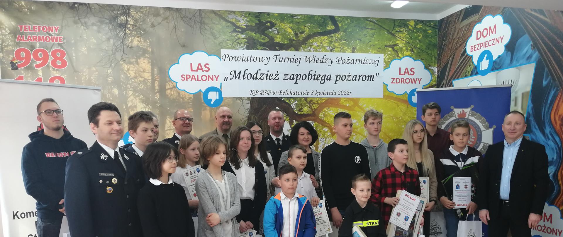 na zdjęciu dzieci nagrodzone w powiatowym turnieju wiedzy pożarniczej, zaproszeni goście oraz komendanci powiatowi państwowej straży pożarnej, w tle plakat turnieju oraz bilbordy straży i ochotniczej straży pożarnej