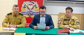 Zdjęcie przedstawia osoby siedzące przy stole z zielonym suknem, na stole leżą notesy oraz czerwona teczka. Po prawej i po lewej stronie funkcjonariusze ubrani w piaskowy mundur służbowy. Po środku Prezes zarządu powiatowego w granatowej marynarce przegląda dokument.