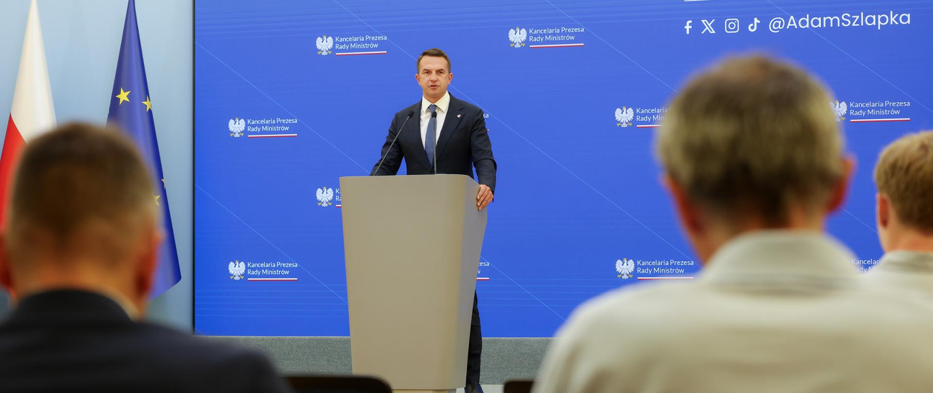 Rzecznik rządu Minister Adam Szłapka podczas konferencji prasowej po posiedzeniu Rady Ministrów 26.08.2025 r. W tle ekran z logo KPRM i kontami Rzecznika w social media.