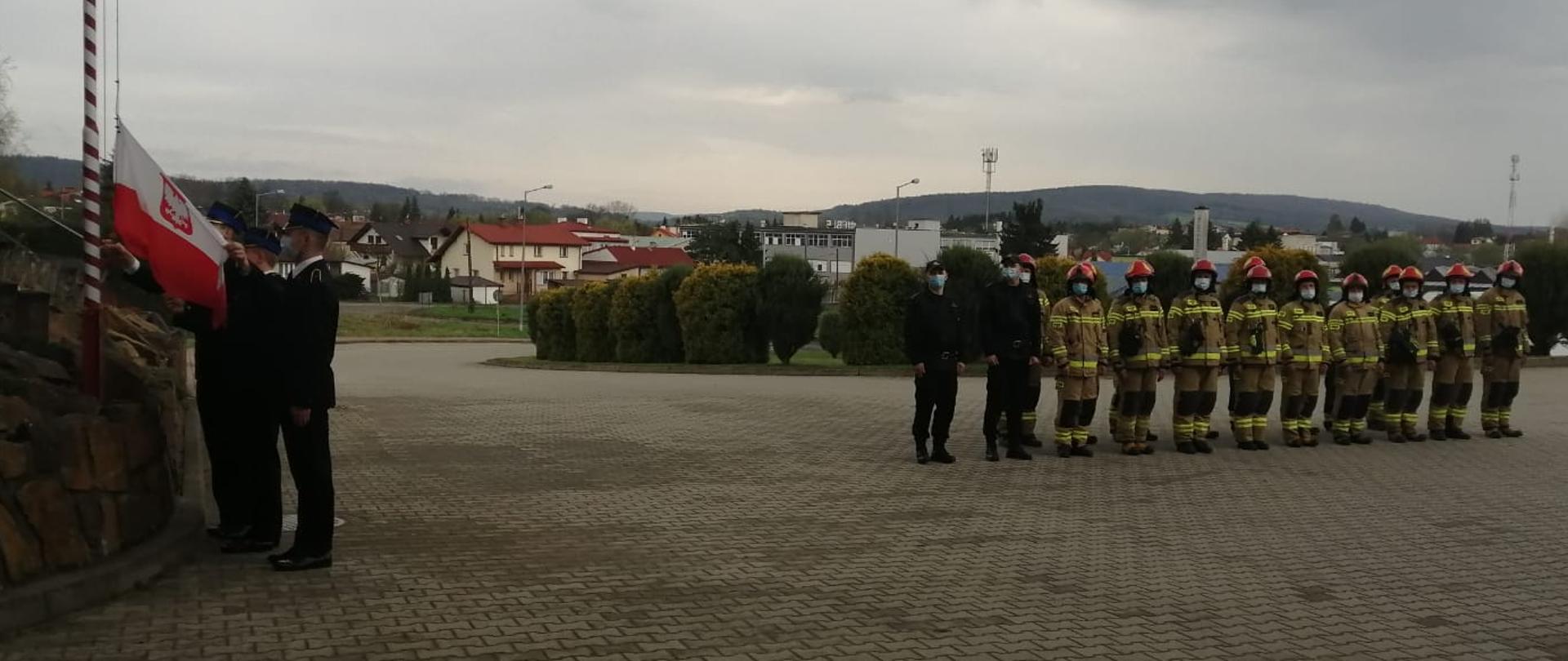 Zdjęcie przedstawia poczet flagowy złożony z 3 strażaków ubranych w umundurowanie wyjściowe. Poczet dokonuje podniesienia flagi narodowej. Po prawej stronie zdjęcia znajduje się 20 osób w umundurowaniu bojowym koloru piaskowego.