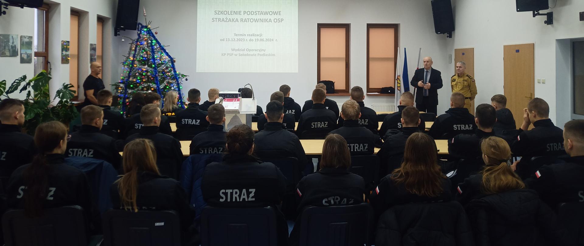 Uczniowie siedzący na szkoleniu strażaka ratownika OSP. W tle osoby prowadzące i świąteczna choinka