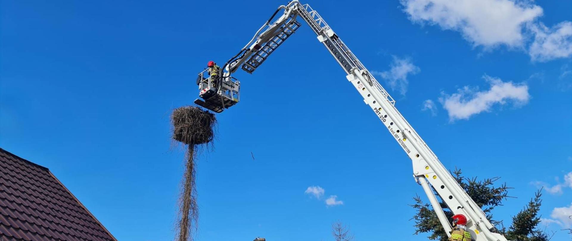 Zdjęcie przedstawia rozstawiony, czerwono - biały strażacki podnośnik hydrauliczny. Znajdujący się na dole strażak w piaskowym mundurze operuje urządzeniem. W koszu, który znajduje się na wysokości strażak ogląda bocianie gniazdo znajdujące się na słupie. W tle błękitne niebo i dachy zabudowań.