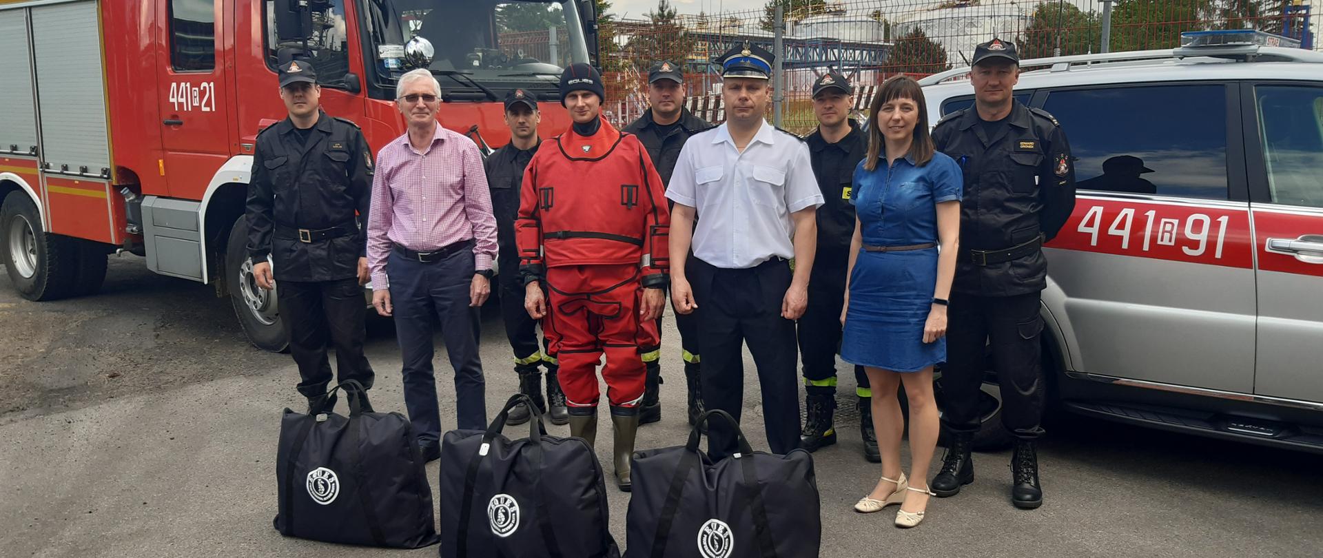 Na zdjęciu stoją kolejno brygadier Jan Szeląg, Prezes Orion Ryszard Rozwadowski, starszy ogniomistrz Mariusz Bara, sekcyjny Damian Gorczyca, kapitan Mateusz Błasik, brygadier Piotr Śmietana komendant powiatowy PSP w Jaśle, starszy strażak Mateusz Kozłecki, Monika Pabisz, młodszy brygadier Edward Gronek. W tle samochody pożarnicze. 