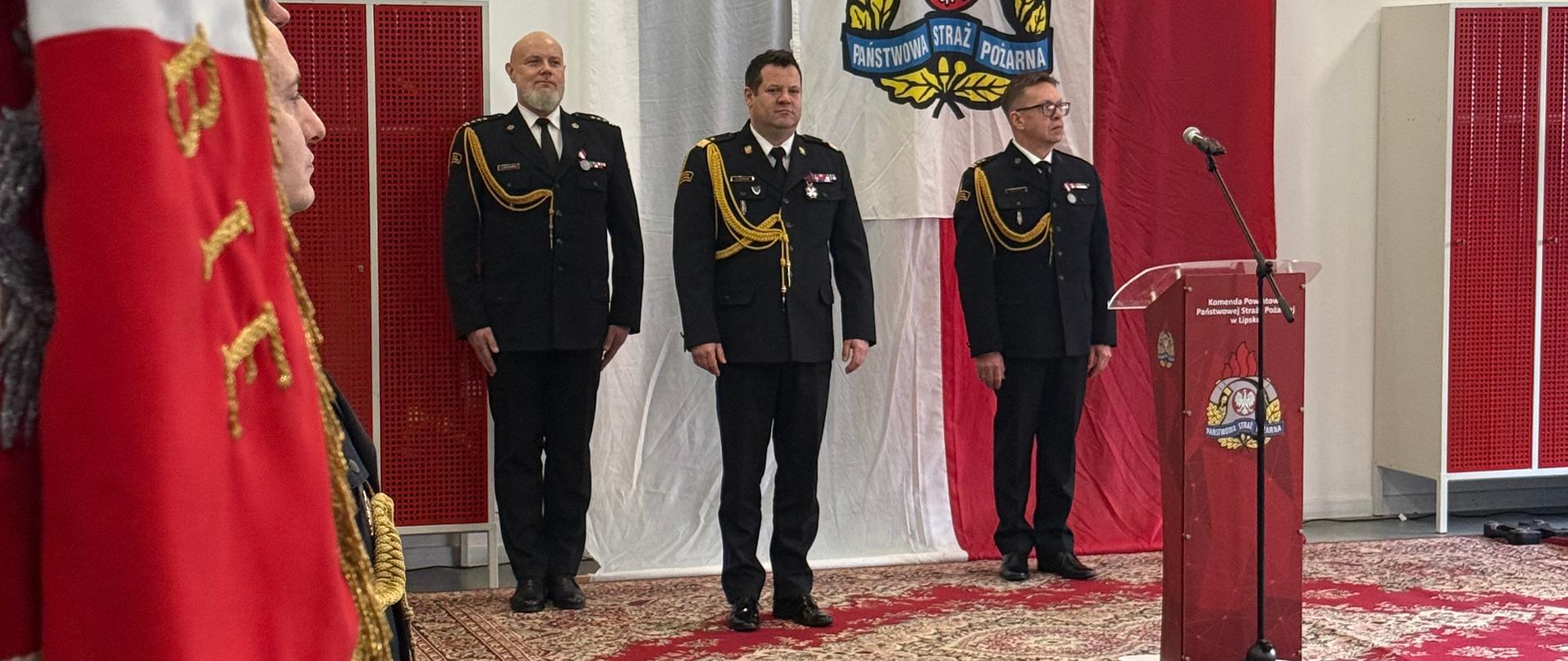 Perspektywa z boku na trzech oficerów straży pożarnej stojących w postawie zasadniczej przy czerwonej mównicy. W tle duża biało-czerwona flaga z godłem PSP na środku. Na pierwszym planie widać fragment materiału sztandaru.