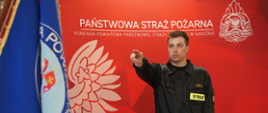 Strażak w czarnym umundurowaniu składa ślubowanie, wyciągając prawą rękę przed siebie podczas uroczystości w Komendzie Powiatowej Państwowej Straży Pożarnej w Gnieźnie. Na pierwszym planie widoczny jest fragment sztandaru, a w tle czerwona ścianka z logo i nazwą Państwowej Straży Pożarnej.