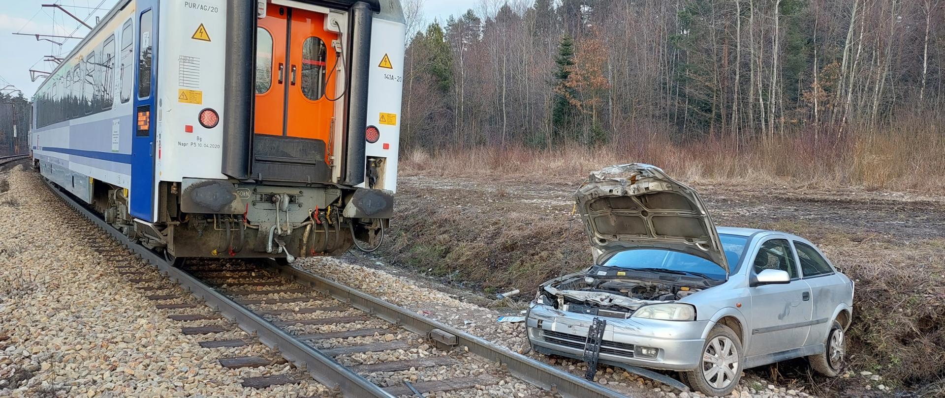 Na zdjęciu przedstawiono: na torze stoi skład osobowy a po jego prawej stronie, w rowie odwadniającym torowisko znajduje się uszkodzony samochód osobowy.