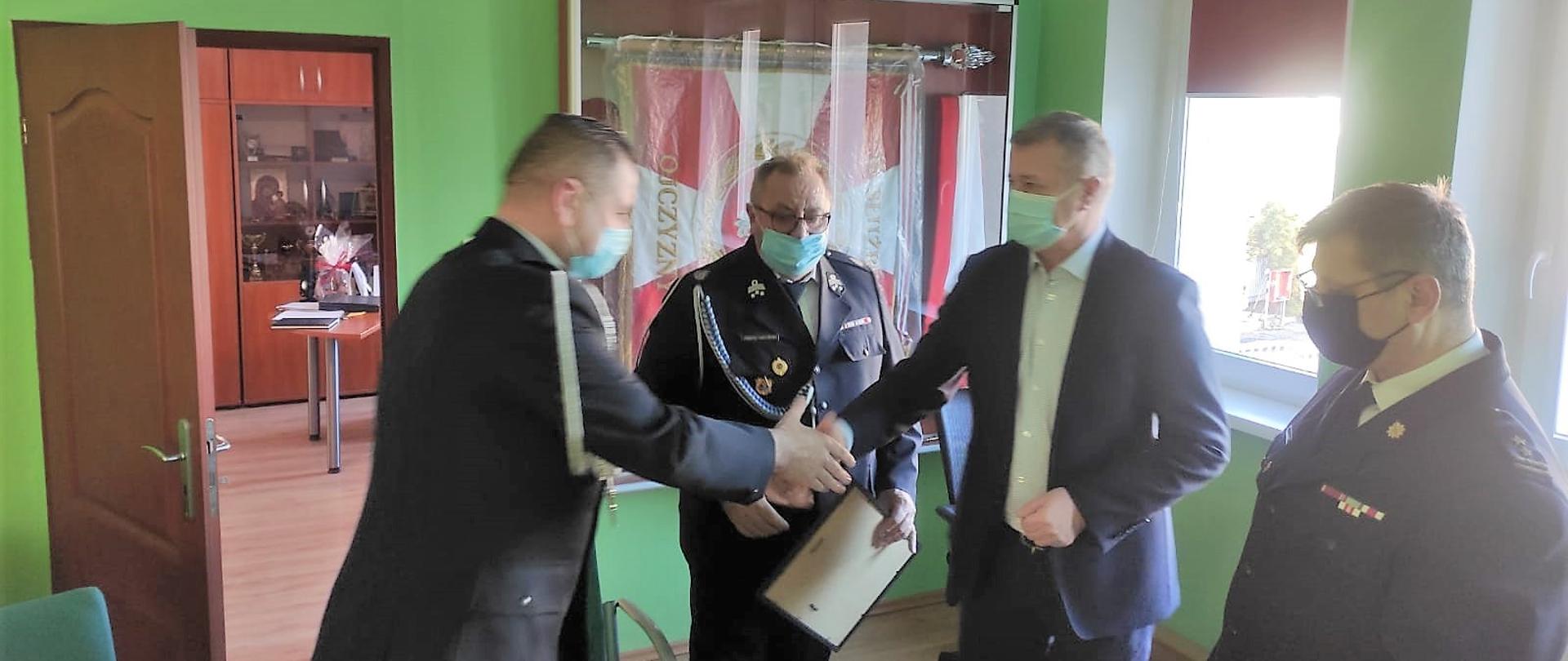 Obraz przedstawia Komendanta Powiatowego brygadiera Waldemara Sarleja, Wójta Gminy Męcinka pana Mirosława Brzozowskiego, Prezesa OSP Małuszów druha Janusza Hołyńskiego oraz Naczelnika OSP Małuszów druha Tomasza Hołyńskiego w trakcie przekazania decyzji o włączeniu jednostki do KSRG. Strażacy w umundurowaniu wyjściowym. Osoby znajdują się w sali narad.