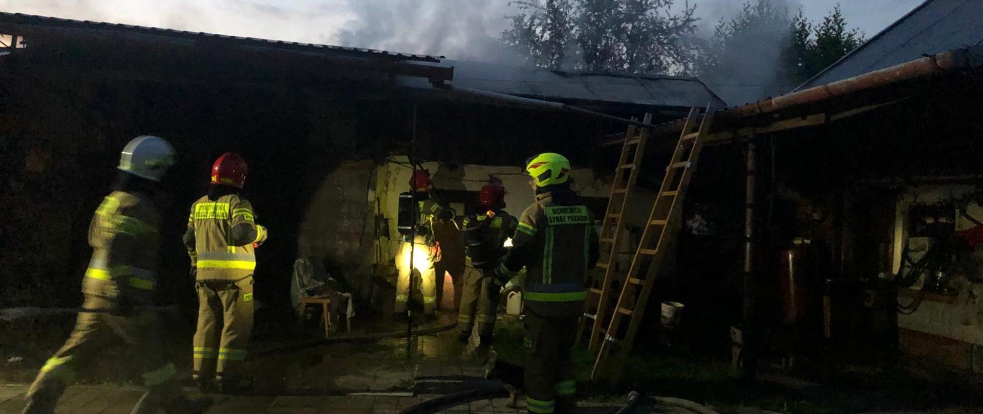 Na zdjęciu widać strażaków podczas gaszenia pożaru budynku gospodarczego, z budynku wydobywa się zadymienie, przed budynkiem linie wężowe .