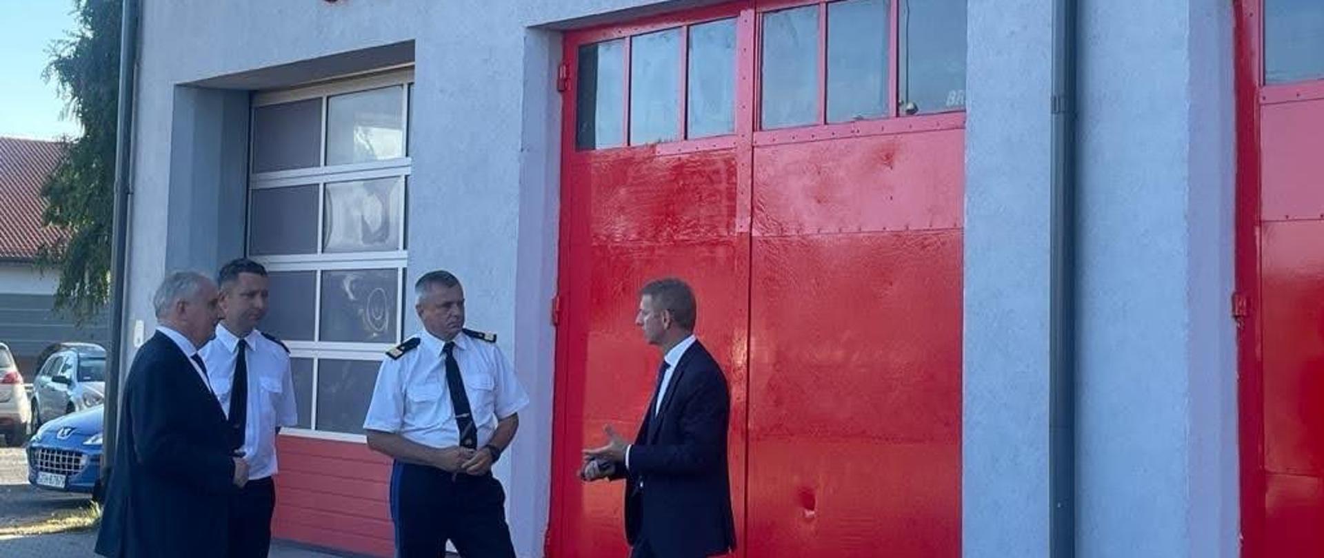Wojewoda pomorski, funkcjonariusze Państwowej Straży Pożarnej oraz przedstawiciel władz gminy stoją przed czerwoną bramą garażową na ścianie jest napis dom strażaka w Brusach. 