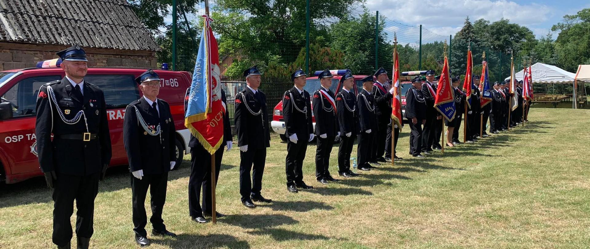 Poczty sztandarowe ustawione w szeregu. W tle samochód pożarniczy