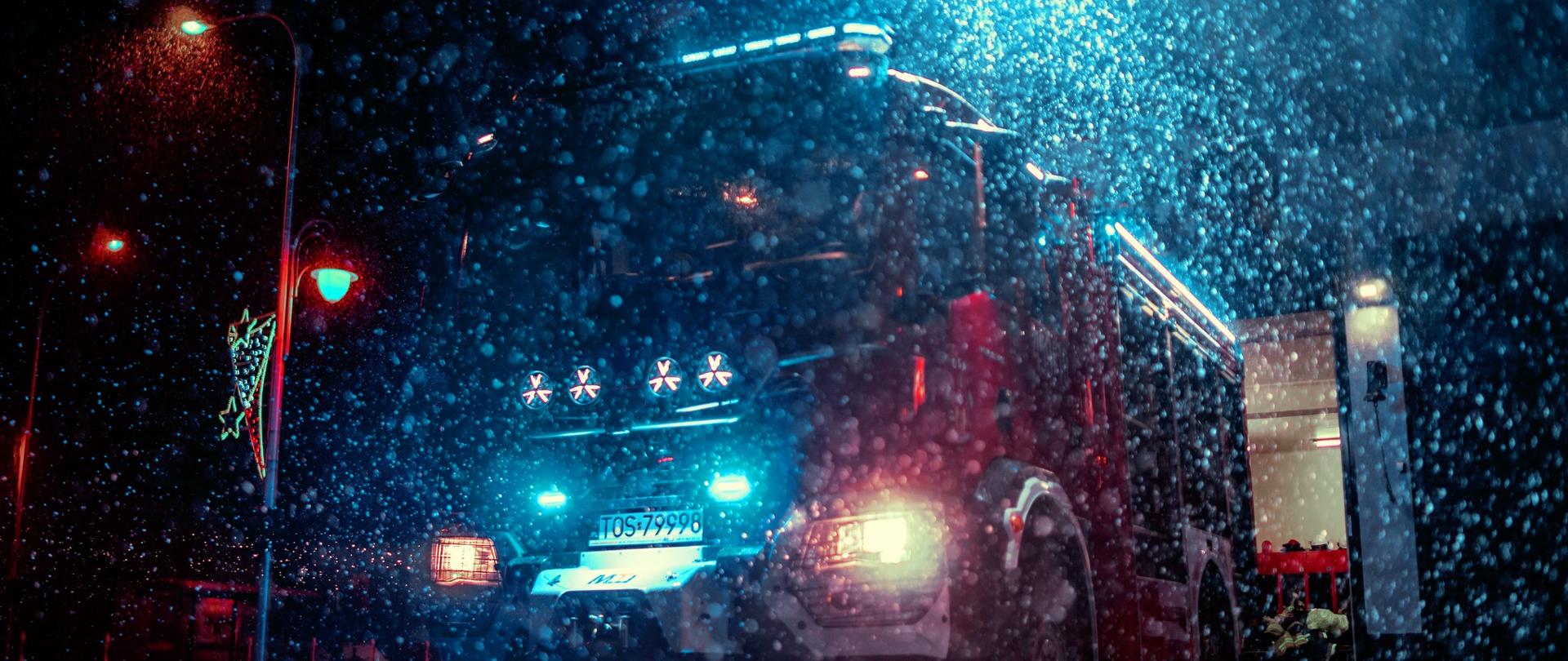 Na zdjęciu w porze nocnej podczas padającego śniegu widać na pierwszym planie oświetlony samochód pożarniczy stojący przodem. 