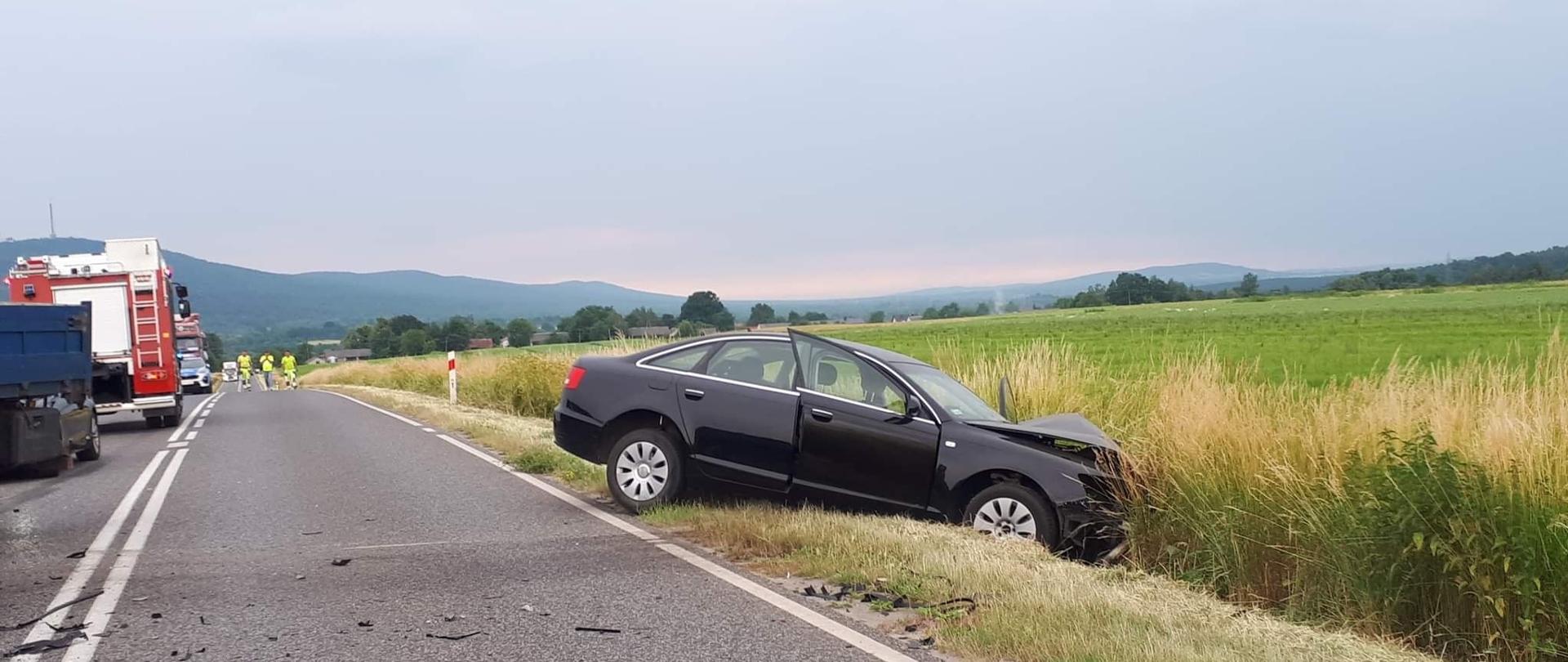 Na zdjęciu widać w przydrożnym rowie samochód osobowy z rozbitym przodem. W oddali widać samochody ratownicze. Zdjęcie wykonane na tle gór świętokrzyskich