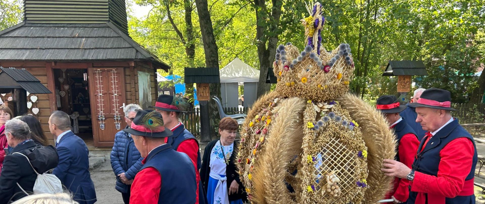 Mężczyźni w tradycyjnych strojach niosą duży, kunsztowny wieniec dożynkowy w kształcie korony, wykonany ze zboża.