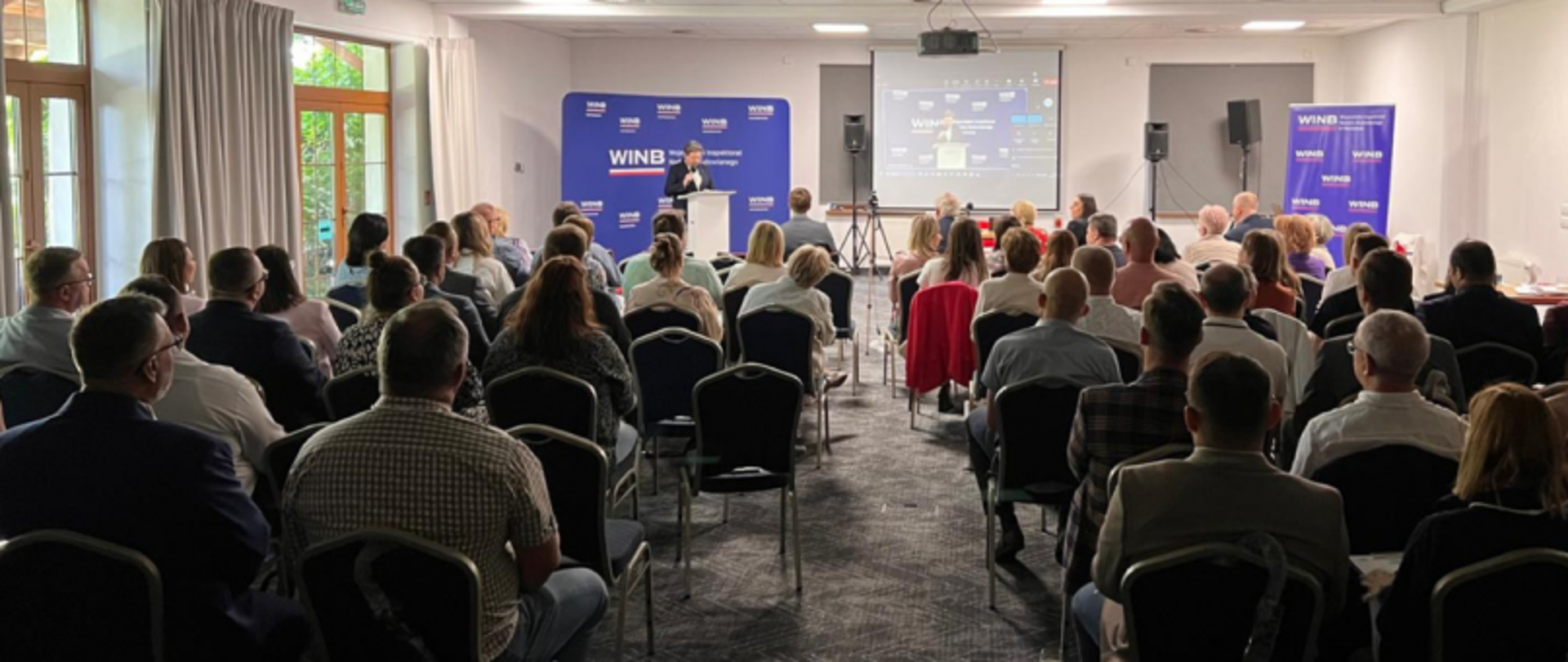 Warsztaty lub szkolenie zorganizowane przez WINB Warszawa – Wojewódzki Inspektorat Nadzoru Budowlanego w Warszawie. Wydarzenie odbywa się w sali konferencyjnej. Widać kilkanaście osób uczestniczących w konferencji.