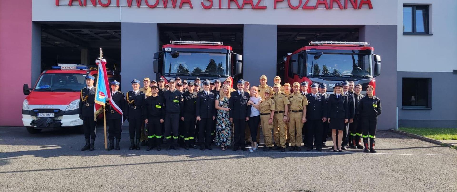 Grupa strażaków stoi w szeregu przed strażnicą. Po lewej stronie jeden z nich trzyma sztandar. W tle trzy samochody strażackie stoją w otwartych bramach garażu. Nad bramami widnieje napis Państwowa Straż Pożarna.