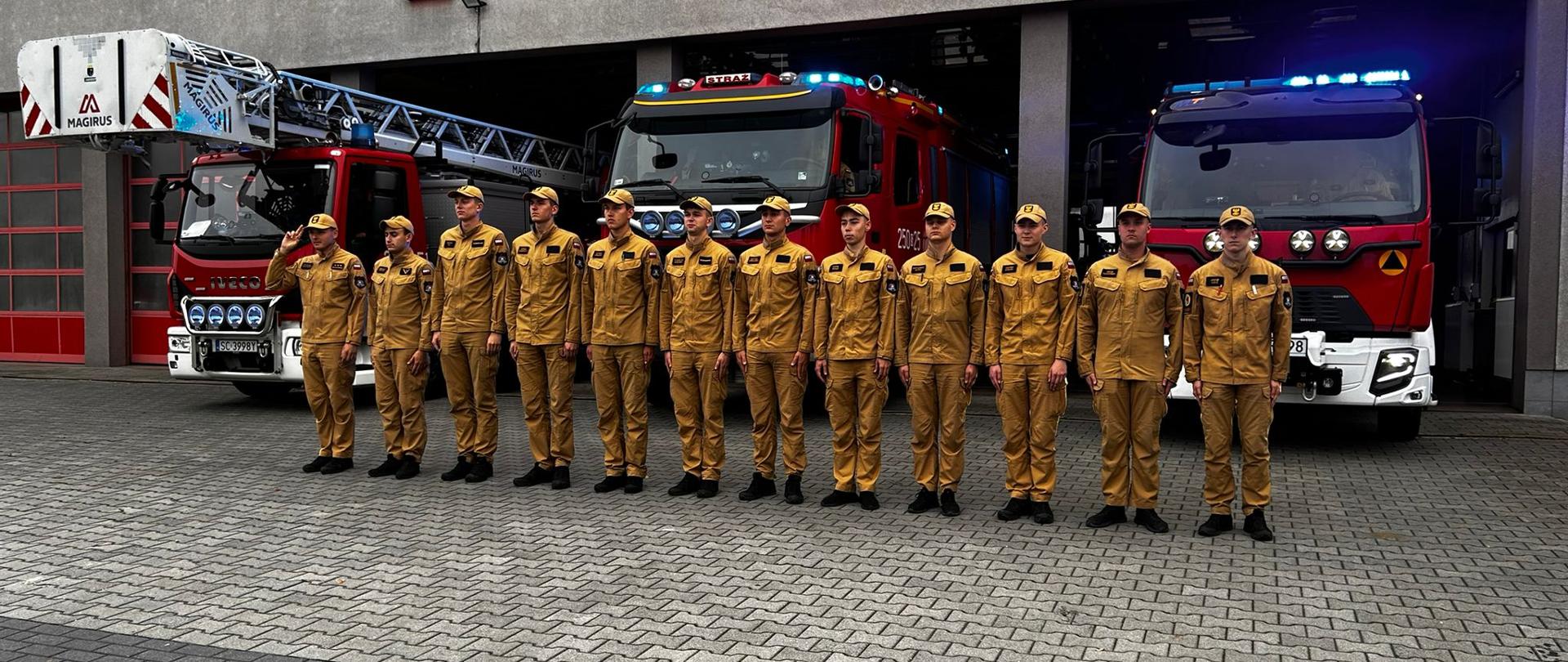 Zastęp strażaków Szkolnej Jednostki Ratowniczo-Gaśniczej stoi przed bramami garażowymi jednostki oddając hołd zmarłemu strażakowi ochotnikowi, w tle stojące samochody bojowe z włączonymi sygnałami świetlnymi