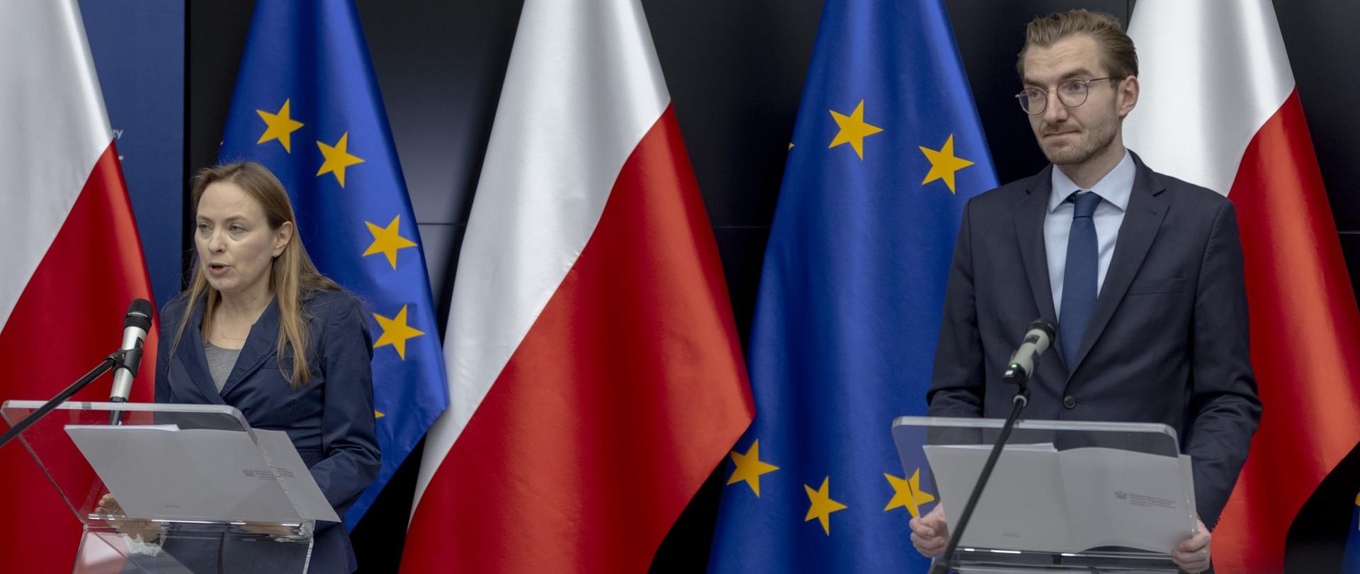 Minister Katarzyna Pełczyńska-Nałęcz oraz wiceminister Jan Szyszko stoją w mównicach przed mikrofonami na tle flag Polski i Unii Europejskiej