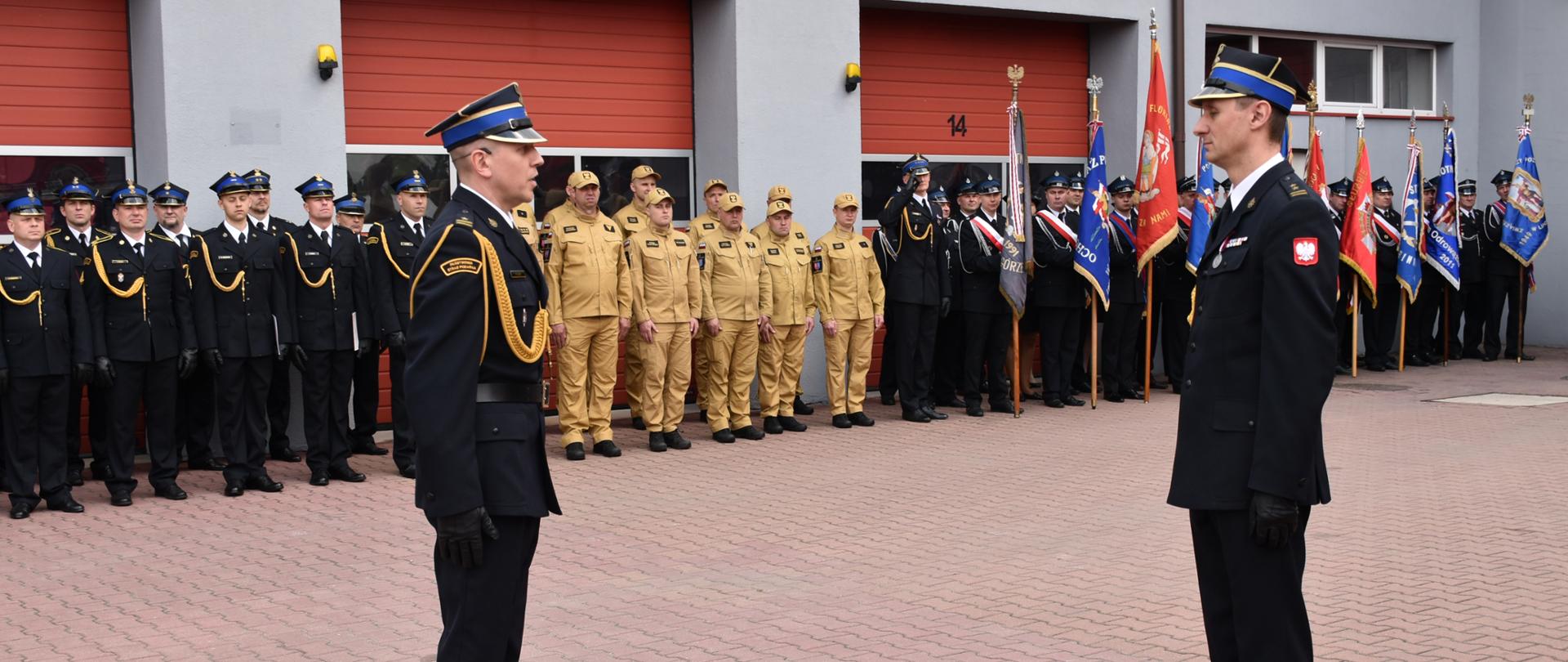 Dwóch oficerów w galowych, ciemnych mundurach stoi naprzeciw siebie na placu przed budynkiem z czerwoną bramą oznaczoną numerem 14. Obaj mają czapki z daszkiem, białe rękawiczki i złote sznury reprezentacyjne. Za nimi w równych rzędach stoją strażacy w czarnych i jasnych mundurach, a po prawej stronie widoczne są poczty sztandarowe z kolorowymi flagami. Scena przedstawia formalny moment uroczystości