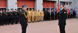 Dwóch oficerów w galowych, ciemnych mundurach stoi naprzeciw siebie na placu przed budynkiem z czerwoną bramą oznaczoną numerem 14. Obaj mają czapki z daszkiem, białe rękawiczki i złote sznury reprezentacyjne. Za nimi w równych rzędach stoją strażacy w czarnych i jasnych mundurach, a po prawej stronie widoczne są poczty sztandarowe z kolorowymi flagami. Scena przedstawia formalny moment uroczystości