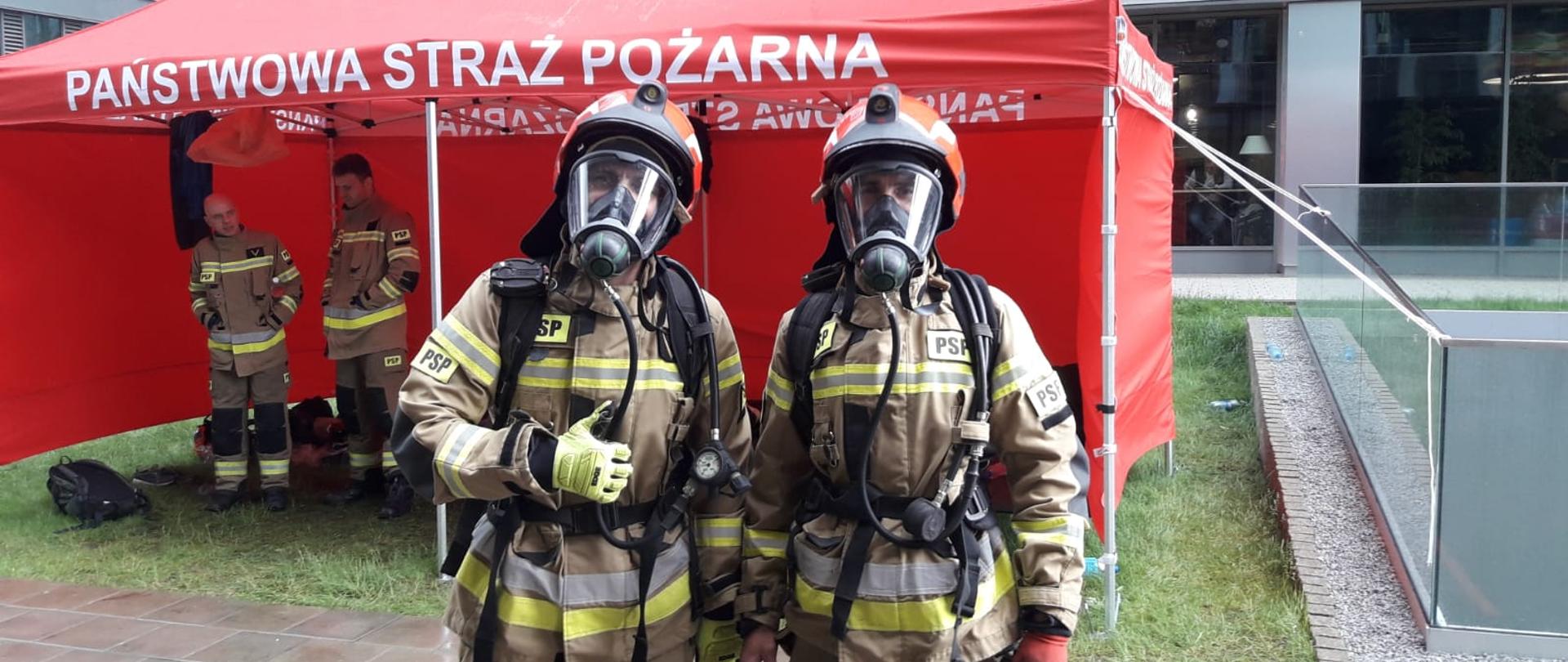 Dwoje strażaków w pełnym umundurowaniu z hełmami na głowie i maskami na twarzy pozuje do zdjęcia. W tle namiot koloru czerwonego. 