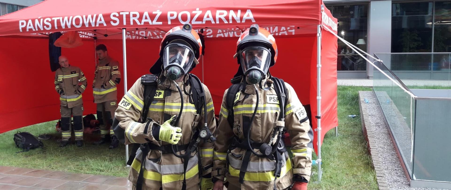 Dwoje strażaków w pełnym umundurowaniu z hełmami na głowie i maskami na twarzy pozuje do zdjęcia. W tle namiot koloru czerwonego. 