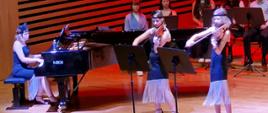 Trzy dziewczynki ubrane w takie same sukienki grają na skrzypcach, altówce i fortepianie
