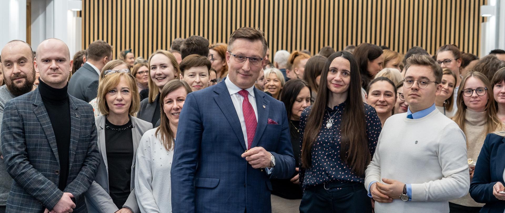 Zdjęcie zbiorowe, na tle ściany z pionowych drewnianych listewek stoi duża grupa osób.