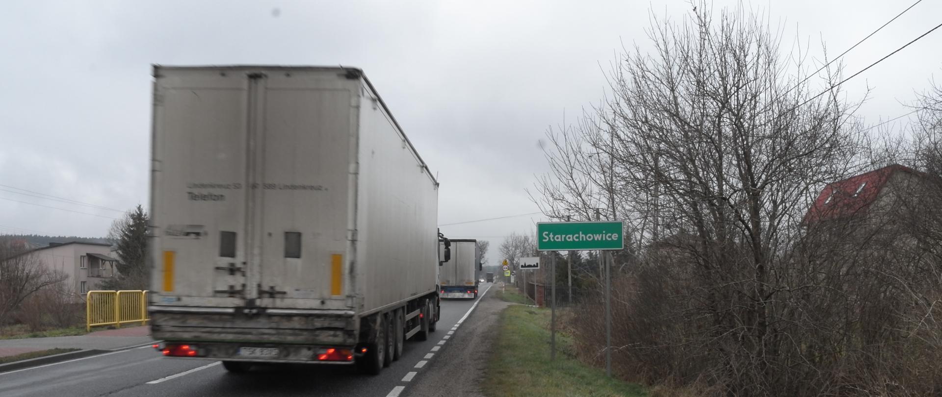 Jednojezdniową drogą krajową jadą samochody ciężarowe jeden za drugim. Przy drodze zielona tablica z nazwą miejscowości Starachowice, dalej tablica oznaczająca teren zabudowany i znaki drogowe. 