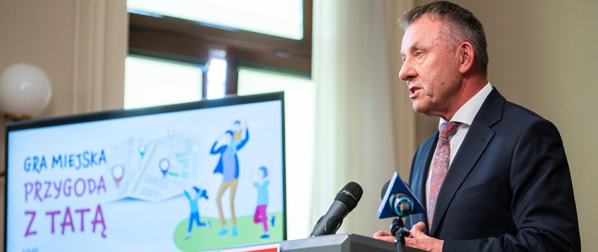 wojewoda łódzki Karol Młynarczyk przemawia przy mównicy, za nim na telewizorze slajd z grafiką promującą grę Przygoda z tatą